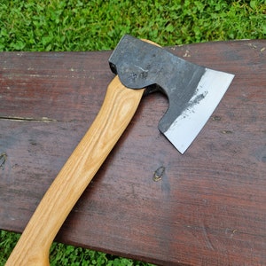 May include: A hand-forged axe with a wooden handle. The axe head is black with a polished edge and has a small mark on the side. The handle is light brown wood.