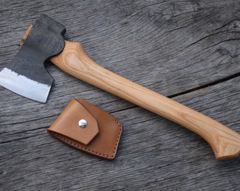 May include: A small, black-handled axe with a polished steel blade and a leather sheath. The axe is lying on a wooden surface.