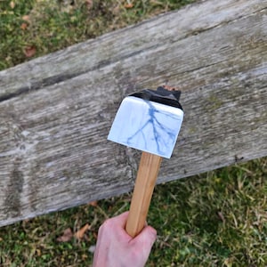May include: An axe with a wooden handle and a silver-coloured head. The axe is being held by a hand wearing a black sleeve.