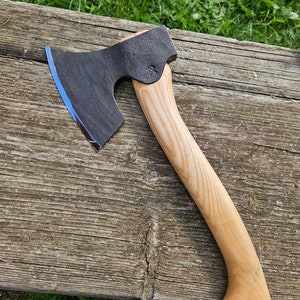 May include: A black axe with a blue edge and a light brown wooden handle. The axe is resting on a wooden surface.