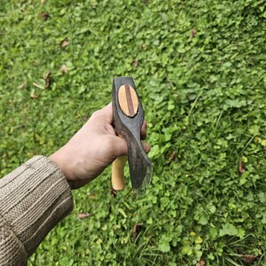 May include: A hand holds a small black axe with a wooden handle. The axe head has a wooden inlay with a striped pattern.
