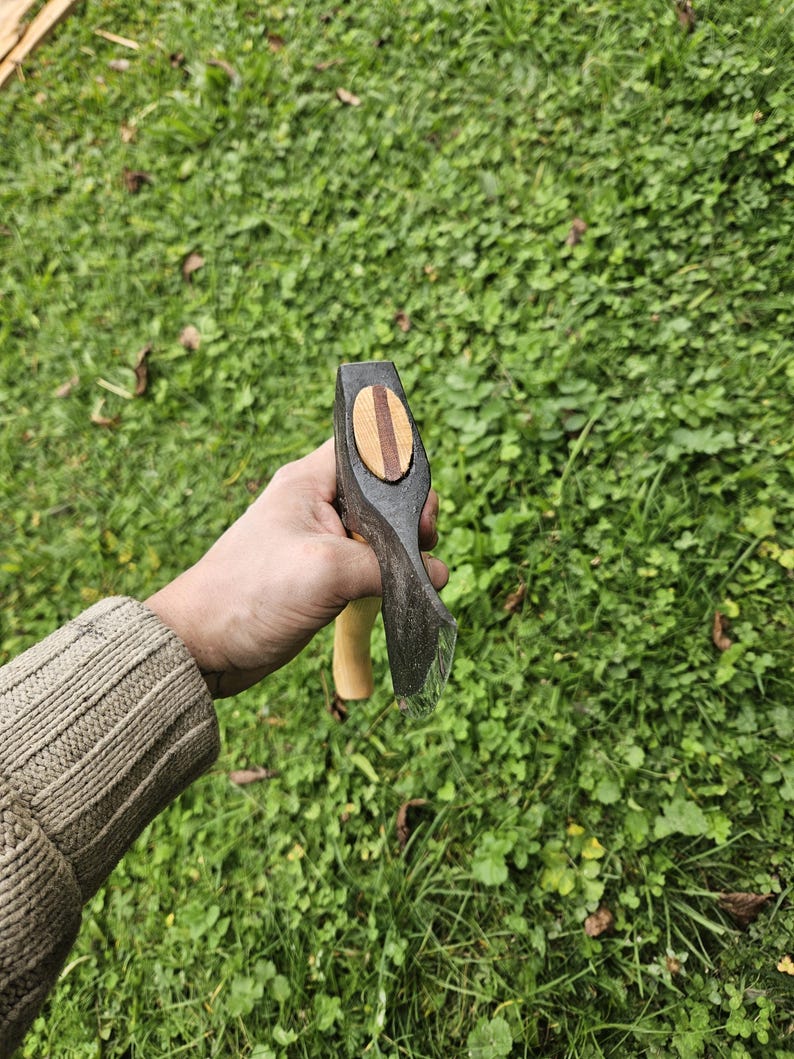May include: A hand holds a small black axe with a wooden handle. The axe head has a wooden inlay with a striped pattern.