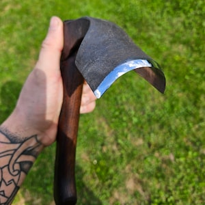 Może przedstawiać: Narzędzie do rzeźbienia z drewnianym uchwytem i zakrzywionym, polerowanym ostrzem ze stali. Narzędzie jest trzymane w dłoni z tatuażem.