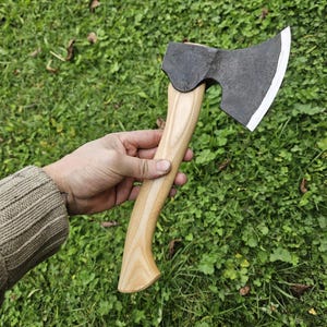 May include: A hand holds a black axe with a wooden handle. The axe has a curved blade and a slightly rounded head. The axe is set against a background of green grass.