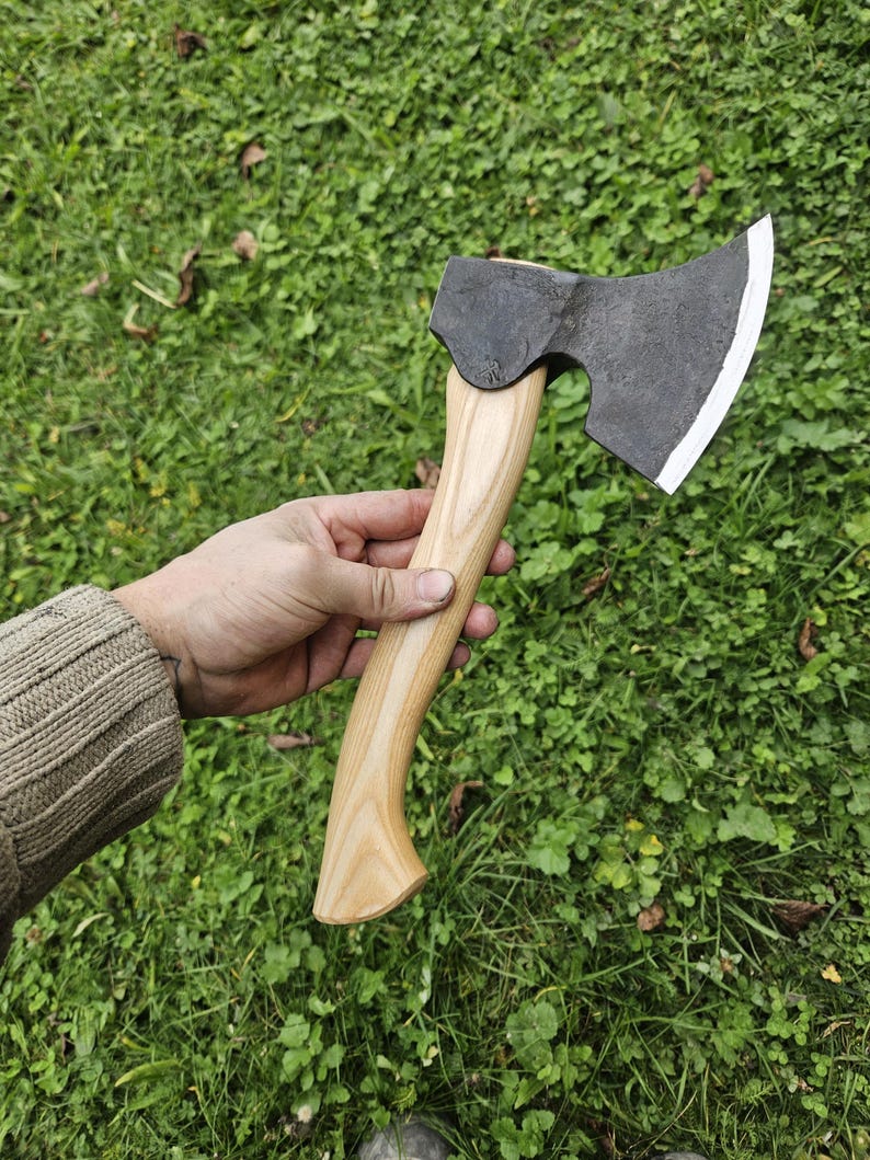 May include: A hand holds a black axe with a wooden handle. The axe has a curved blade and a slightly rounded head. The axe is set against a background of green grass.