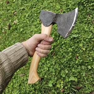 May include: A hand holds a black axe with a wooden handle. The axe has a curved blade and a slightly rounded head. The axe is held against a background of green grass.