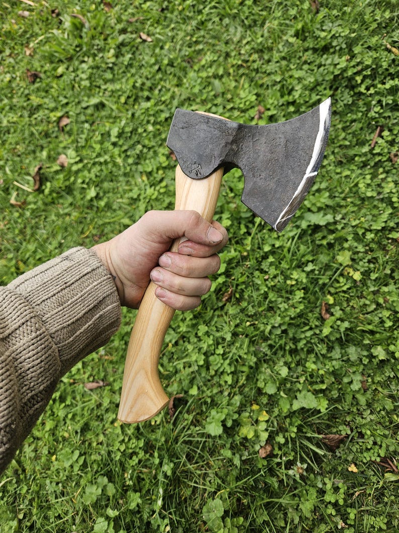 May include: A hand holds a black axe with a wooden handle. The axe has a curved blade and a slightly rounded head. The axe is held against a background of green grass.