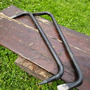 May include: Two dark gray steel pry bars with curved handles and flat, beveled ends. One bar has a pointed end. The tools are resting on a weathered, brown wooden surface, with green grass in the background.