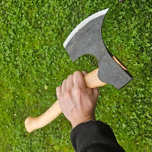 Może przedstawiać: Ręka trzyma czarną siekierę z drewnianym trzonkiem. Siekiera ma zakrzywione ostrze i zaokrągloną głowicę. Siekiera znajduje się na zielonym tle trawy.