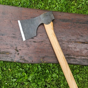 Könnte beinhalten: Eine schwarze Axt mit einem Holzstiel. Der Axtkopf hat eine gebogene Klinge und einen kleinen, abgerundeten Hammer auf der Rückseite. Der Stiel ist aus hellem Holz gefertigt und hat eine glatte Oberfläche.