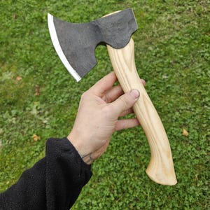 May include: A hand holds a small black axe with a wooden handle. The axe has a curved blade and a slightly rounded head. The handle is light brown and has a smooth finish.