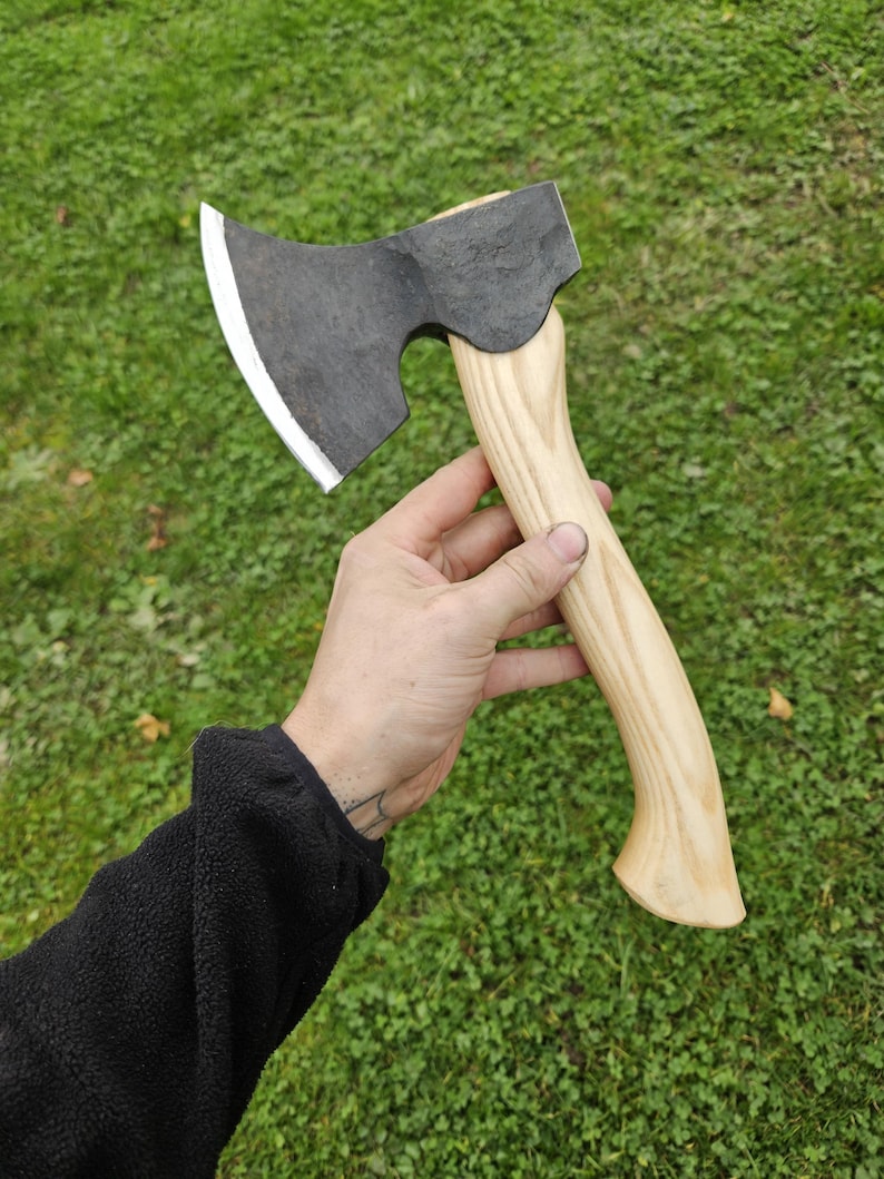 May include: A hand holds a small black axe with a wooden handle. The axe has a curved blade and a slightly rounded head. The handle is light brown and has a smooth finish.