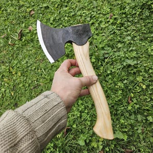 May include: A black hand axe with a wooden handle. The axe has a curved blade and a slightly rounded head. The handle is made of light wood and has a natural finish.