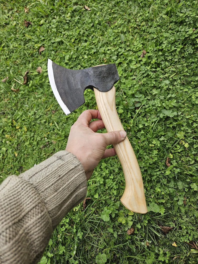 May include: A black hand axe with a wooden handle. The axe has a curved blade and a slightly rounded head. The handle is made of light wood and has a natural finish.