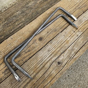 May include: Two metal hooks with pointed ends, lying on a wooden surface. The hooks are made of a dark metal and have a simple, utilitarian design.