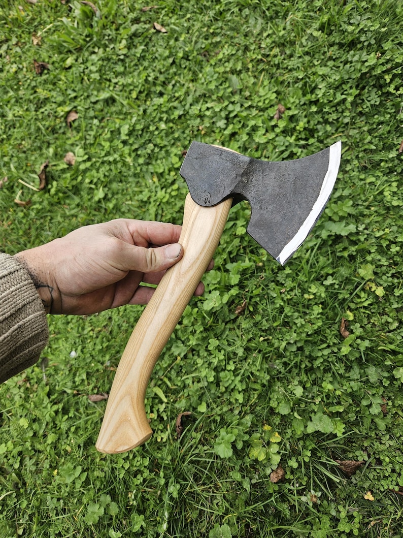 May include: A hand holds a black axe with a wooden handle. The axe has a curved blade and a slightly rounded head. The axe is set against a background of green grass.