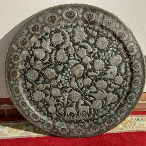 May include: A large, round, antique metal tray with intricate floral and animal engravings. The tray features a detailed pattern of flowers, birds, and deer in a dark silver colour, with a decorative border. The tray is likely used for serving or display.