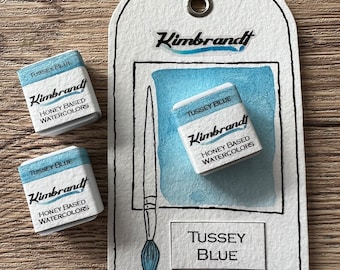 May include: Three watercolor paint pans labeled "Tussey Blue" and "Kimbrandt Honey Based Watercolors". The pans are on a wooden surface with a white tag that says "Tussey Blue" and "Kimbrandt" in black lettering.