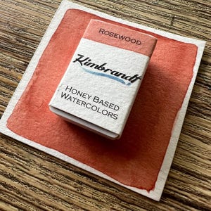 May include: A watercolor paint set labeled "Rosewood" by Kimbrandt, resting on a painted swatch of the same color. The paint set is white with the brand name and "Honey Based Watercolors" printed on it. The background is a wooden surface.