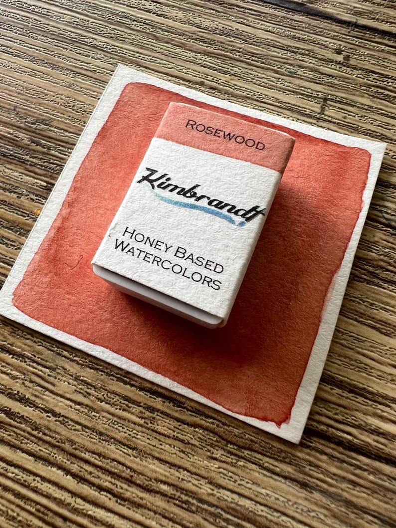 May include: A watercolor paint set labeled "Rosewood" by Kimbrandt, resting on a painted swatch of the same color. The paint set is white with the brand name and "Honey Based Watercolors" printed on it. The background is a wooden surface.