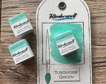 May include: A close-up of watercolor paints and a tag. The tag has a drawing of a paintbrush and a swatch of turquoise green paint. The tag reads "Turquoise Green" and "Kimbrandt Honey Based Watercolors". Two additional paint cubes are also visible.