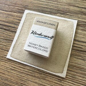 May include: A rectangular box of "Vintage Linen" honey-based watercolors by Kimbrandt, resting on a watercolor swatch. The box is white with black text and a blue accent. The swatch is a light brown color, and the background is a wooden surface.