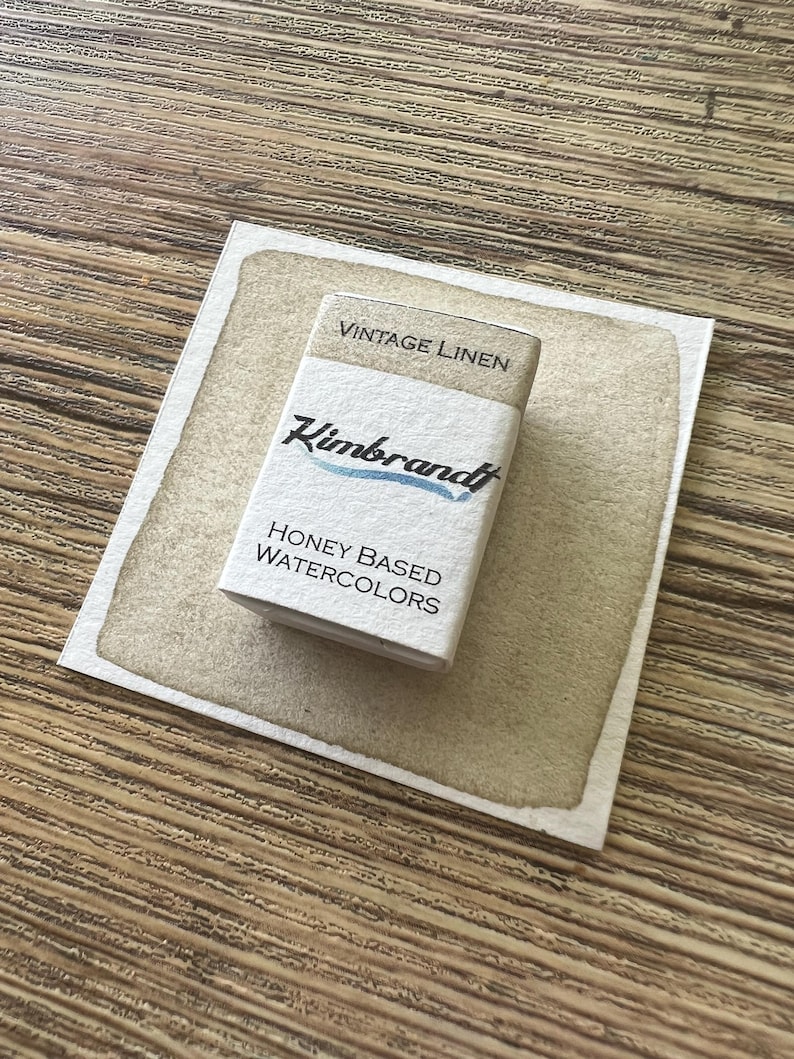 May include: A rectangular box of "Vintage Linen" honey-based watercolors by Kimbrandt, resting on a watercolor swatch. The box is white with black text and a blue accent. The swatch is a light brown color, and the background is a wooden surface.