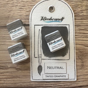 May include: A watercolor art set featuring three neutral tinted graphite honey-based watercolor cubes. The set includes a tag with a brush illustration and the words "Neutral" and "Tinted Graphite". The brand name "Kimbrandt" is visible.