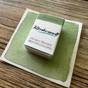 May include: A small box of "Pistachio" honey-based watercolors by "Kimbrandt" sits on a light green watercolor swatch. The box is white with green accents and the brand name in blue. The background is a wooden surface.