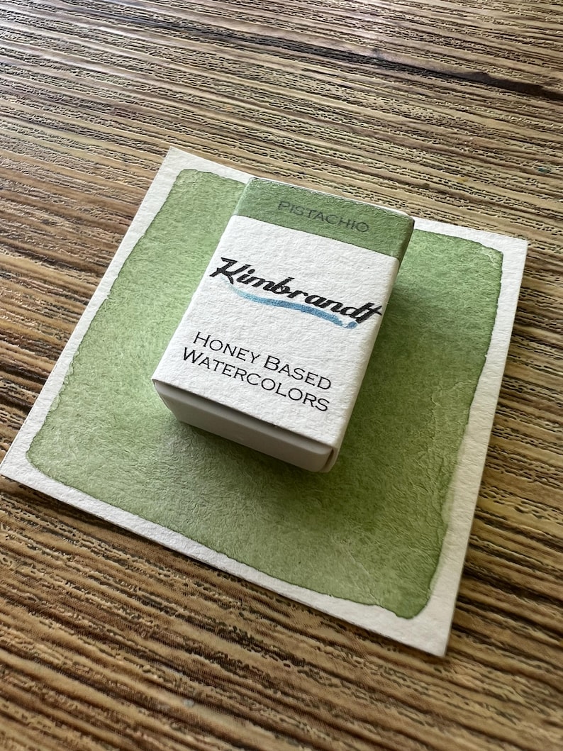 May include: A small box of "Pistachio" honey-based watercolors by "Kimbrandt" sits on a light green watercolor swatch. The box is white with green accents and the brand name in blue. The background is a wooden surface.