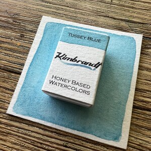 May include: A rectangular watercolor paint set labeled "Tussey Blue" by Kimbrandt, resting on a light blue watercolor swatch. The set is white with black text, and the swatch has a textured, uneven edge. The words "Honey Based Watercolors" are also visible.