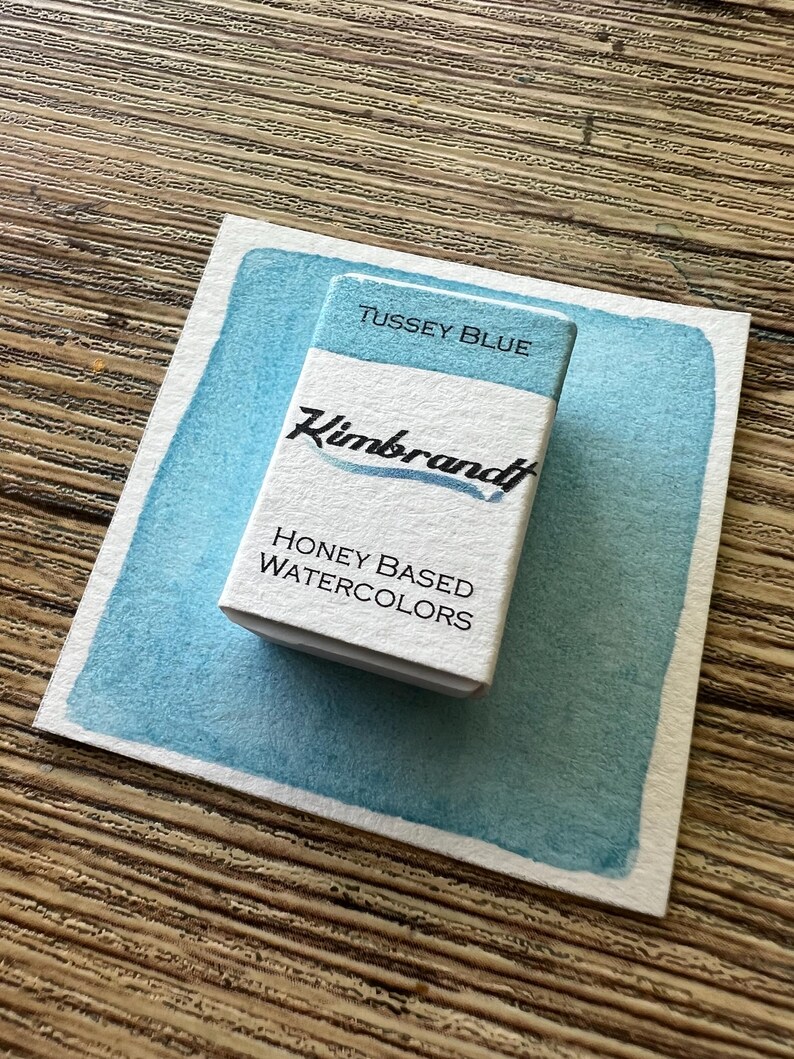 May include: A rectangular watercolor paint set labeled "Tussey Blue" by Kimbrandt, resting on a light blue watercolor swatch. The set is white with black text, and the swatch has a textured, uneven edge. The words "Honey Based Watercolors" are also visible.