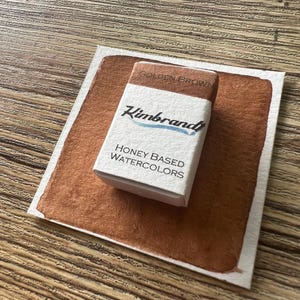 May include: A watercolor paint swatch and a small box of "Kimbrand" honey-based watercolors. The swatch is a rich golden brown color, and the box is white with black and blue text. The box reads "Golden Brown" on top.