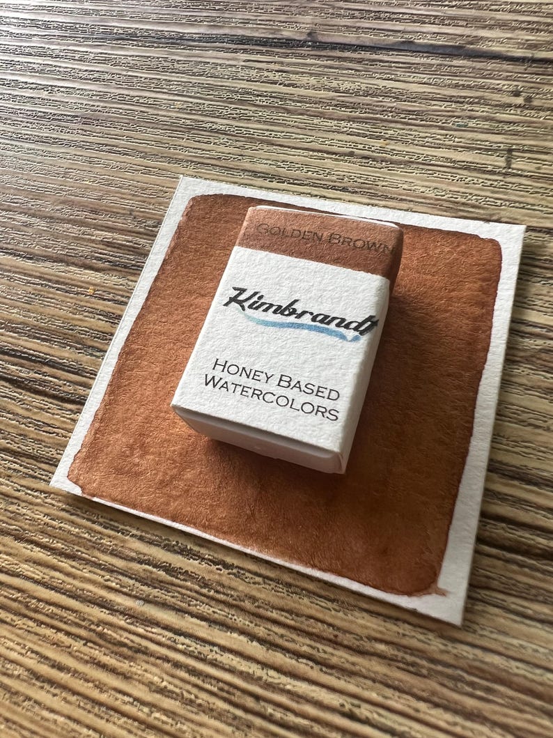 May include: A watercolor paint swatch and a small box of "Kimbrand" honey-based watercolors. The swatch is a rich golden brown color, and the box is white with black and blue text. The box reads "Golden Brown" on top.