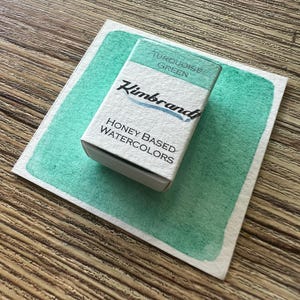 May include: A rectangular box of turquoise green watercolor paint sits on a painted swatch of the same color. The box is labeled "Turquoise Green" and "Honey Based Watercolors" with the brand name "Kimbrand" in a script font.