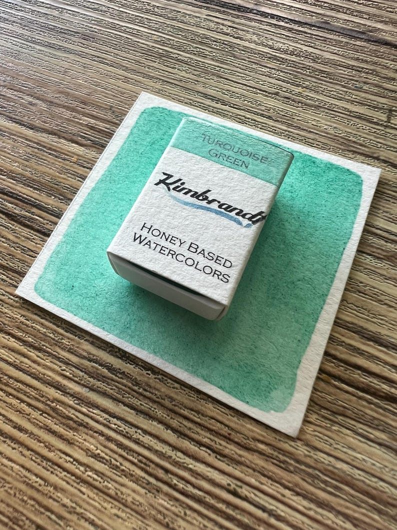 May include: A rectangular box of turquoise green watercolor paint sits on a painted swatch of the same color. The box is labeled "Turquoise Green" and "Honey Based Watercolors" with the brand name "Kimbrand" in a script font.