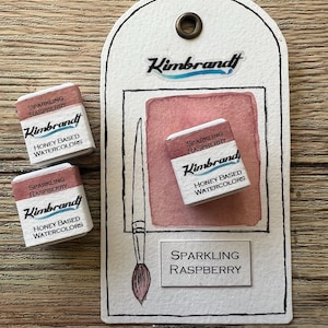 May include: Three watercolour paint pans in a set. The pans are labelled "Sparkling Raspberry" and "Kimbrandt Honey Based Watercolours". The pans are on a wooden surface with a paintbrush and a tag that says "Sparkling Raspberry" and "Kimbrandt".