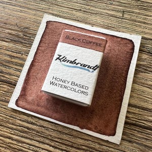 May include: A watercolor paint swatch with a rectangular block of "Black Coffee" honey-based watercolors by Kimbrandt. The swatch is a deep brown color with a white border, and the paint block is white with black and blue text.