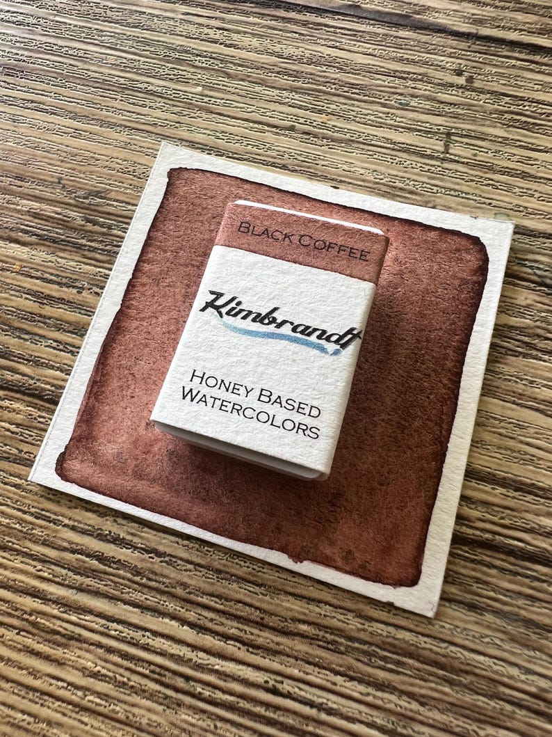 May include: A watercolor paint swatch with a rectangular block of "Black Coffee" honey-based watercolors by Kimbrandt. The swatch is a deep brown color with a white border, and the paint block is white with black and blue text.