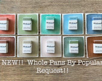 May include: A collection of ten watercolor paint pans, each in a different color, displayed on a wooden surface. Each pan is in a white box with the brand name "Rembrandt" and the text "Honey Based Watercolors". The background colors vary from red to blue.