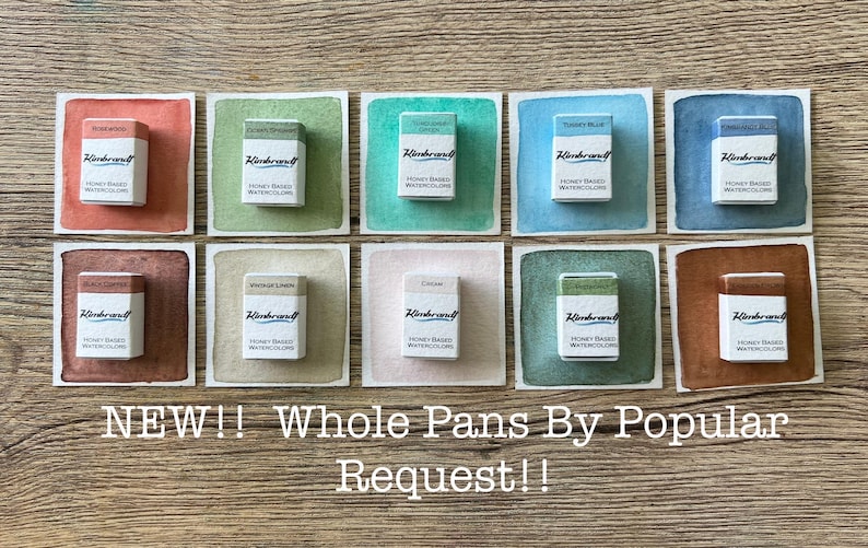 May include: A collection of ten watercolor paint pans, each in a different color, displayed on a wooden surface. Each pan is in a white box with the brand name "Rembrandt" and the text "Honey Based Watercolors". The background colors vary from red to blue.