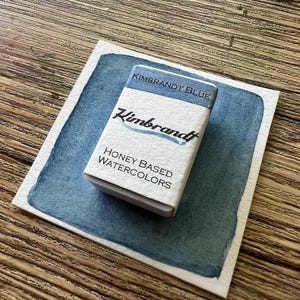 May include: A small white box of "Kimbrandt Blue" honey-based watercolors sits on a blue watercolor swatch. The box has the brand name and product information printed on it. The background is a wooden surface.