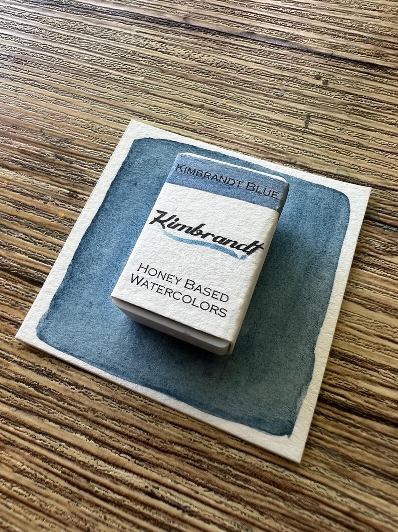 May include: A small white box of "Kimbrandt Blue" honey-based watercolors sits on a blue watercolor swatch. The box has the brand name and product information printed on it. The background is a wooden surface.