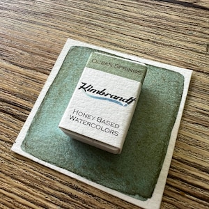 May include: A small box of "Kimbrandt" honey-based watercolors, labeled "Ocean Springs", sits on a watercolor swatch. The box is white with green and blue text. The watercolor swatch is a square of paper with a green wash.