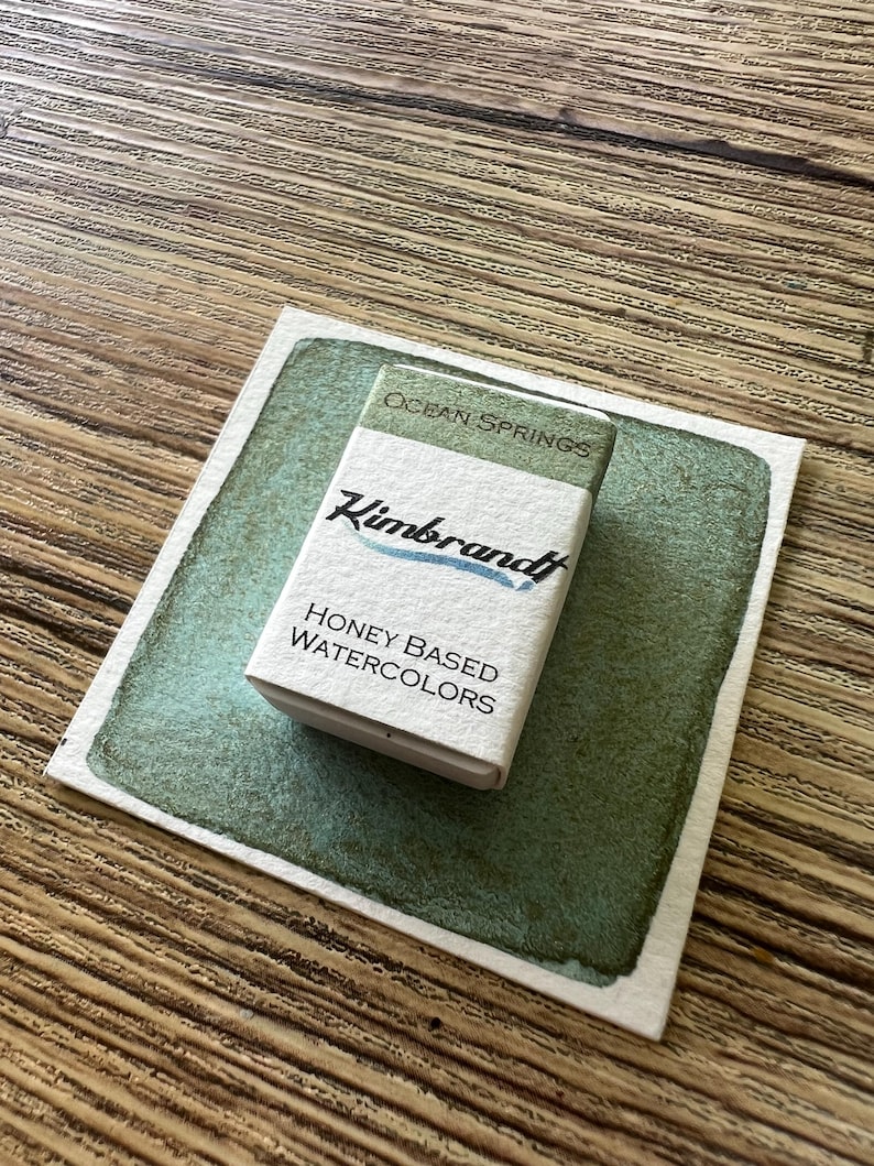 May include: A small box of "Kimbrandt" honey-based watercolors, labeled "Ocean Springs", sits on a watercolor swatch. The box is white with green and blue text. The watercolor swatch is a square of paper with a green wash.