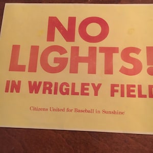 May include: A yellow sign with red text that reads "NO LIGHTS! IN WRIGLEY FIELD" and the tagline "Citizens United for Baseball in Sunshine".