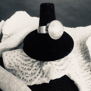 May include: A silver ring with a large, round, white pearl set in a bezel setting. The ring is displayed on a black velvet ring stand.