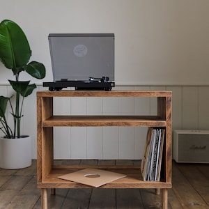 May include: Wooden record player stand with a black turntable. The stand has two shelves, one with vinyl records and the other with a record sleeve. The stand is made of light brown wood and has four short legs. The unit is 75cm high.