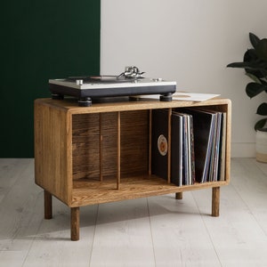 May include: A wooden record player stand with a turntable on top and vinyl records in the shelves. The stand has a dark wood finish and is sitting on a light wood floor.