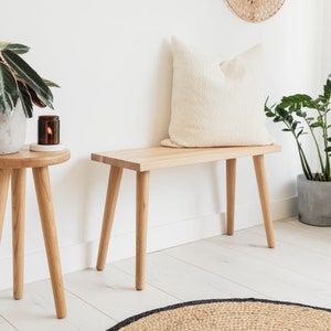 May include: A light wood bench with a white cushion and three legs. The bench is in a room with white walls and a light wood floor. There is a small table with a candle on it in the foreground.